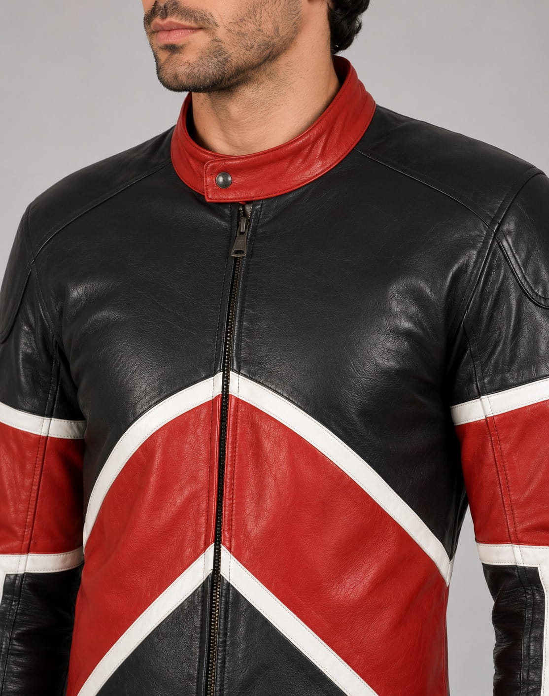 Close-up front view of a male model with dark wavy hair and light stubble wearing a color-blocked racing-style leather jumpsuit featuring black, white, and red paneling with a collarless zip-front finish against a light beige studio backdrop.