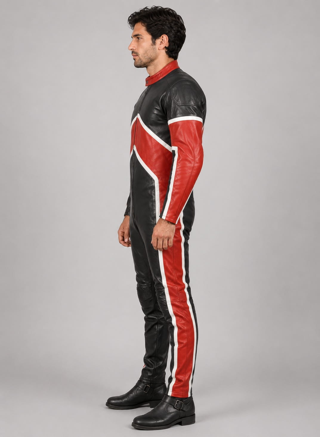 side up view of a male model with dark wavy hair and light stubble wearing a color-blocked racing-style leather jumpsuit featuring black, white, and red paneling with a collarless zip-front finish against a light beige studio backdrop.