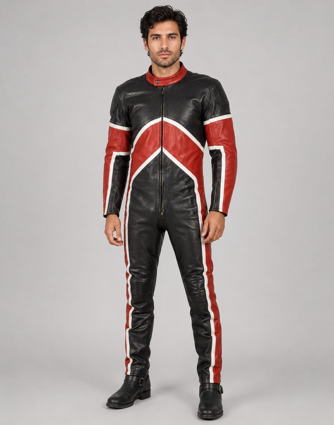 Full-length front view of a male model modeling a fitted black, white, and red leather biker jumpsuit with star accents on the sleeves and ankles, paired with brown leather boots against a solid neutral backdrop.